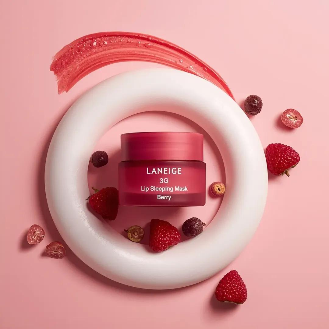 一个粉色化妆品罐居于画面中央,标签为“LANEIGE Lip Sleeping Mask [Berry] 3 g”,放置在一大团旋涡状白色奶油…, 图片 1