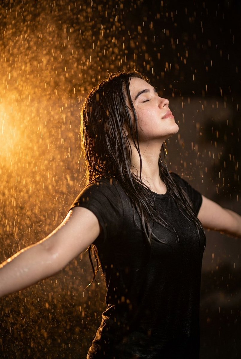 超写实电影感人像摄影:一位32岁的年轻女性在夜晚的暴雨中站立,双臂张开,面部仰向天空,双眼紧闭,感受着雨水。戏剧性的暖橙色背光照亮了坠落的雨滴…, 图片 1