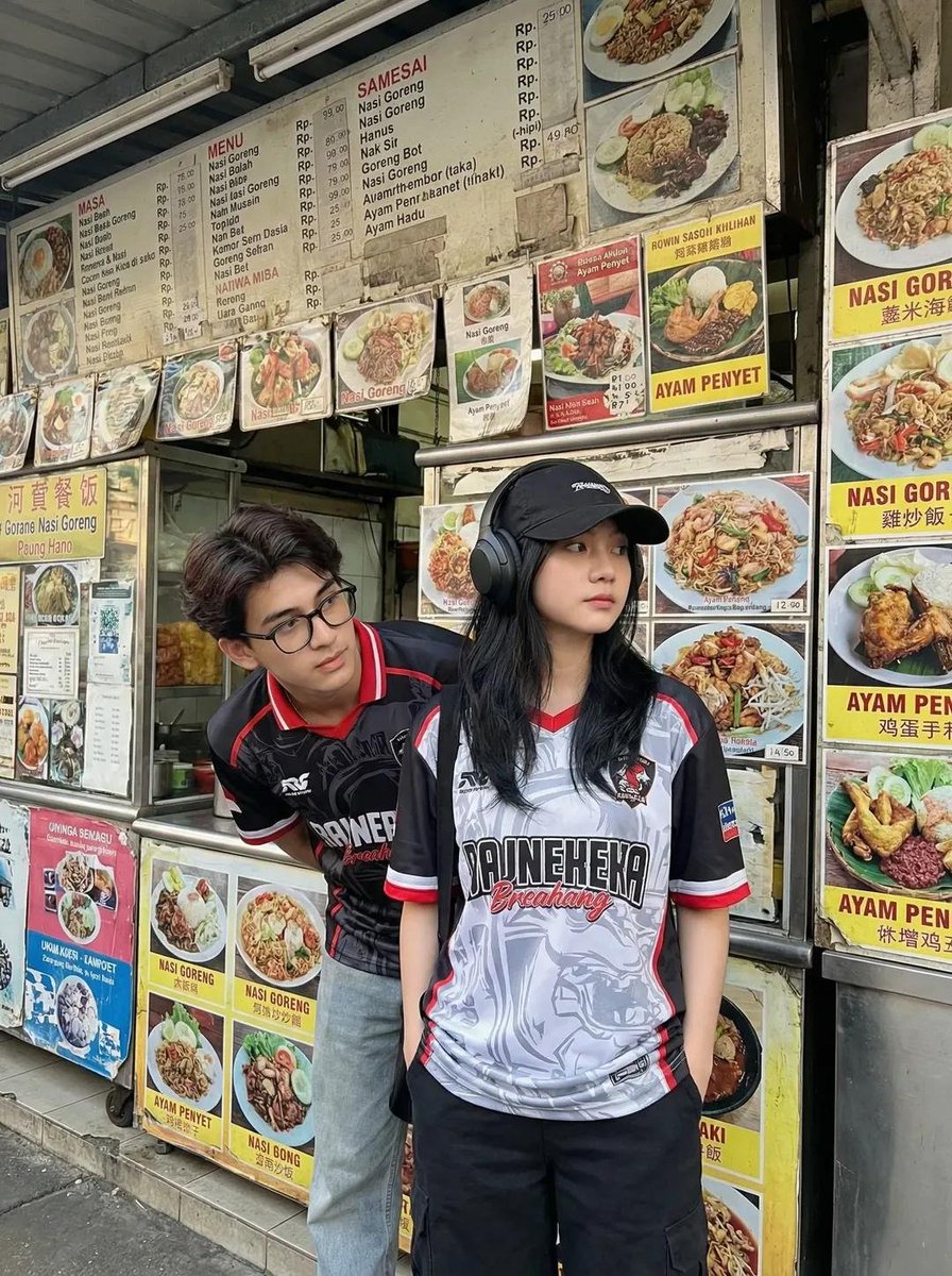 一张抓拍风格的街拍照片:两位年轻的亚洲成年人站在一家小型东南亚街边小吃摊前。小吃摊上挂满了色彩鲜艳的菜单板和食物照片,主打菜品包括 nasi …, 图片 1