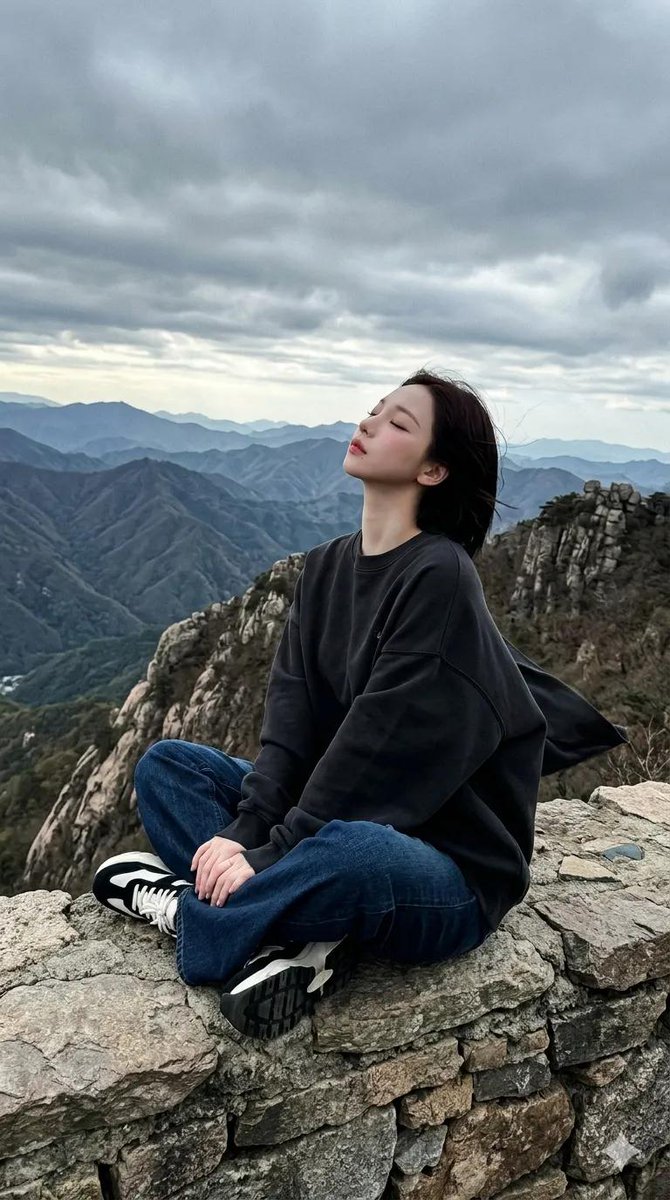 一段电影般的全景画面在戏剧性阴云笼罩的天空下，于一处嶙峋的山脊上徐徐展开。一位女性盘腿坐于石质岩架之上，身着超大号白色印花卫衣、深色阔腿牛仔裤…, 图片 1