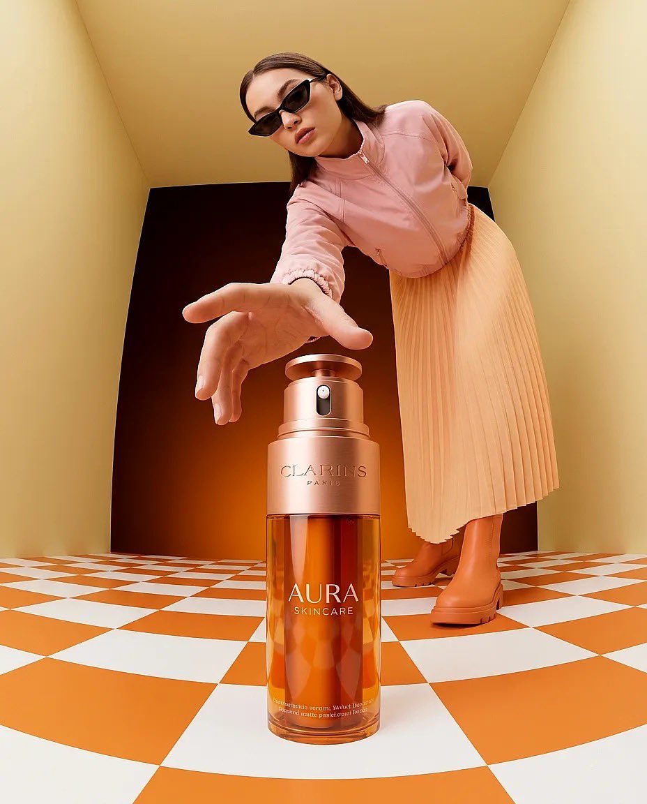 “超现实时尚产品摄影：一只巨大的琥珀色护肤品瓶（Clarins Aura）放置在一个极简盒子房间内的橙白格子地板上，温暖的纯橙色灯光，一位时尚…, 图片 1