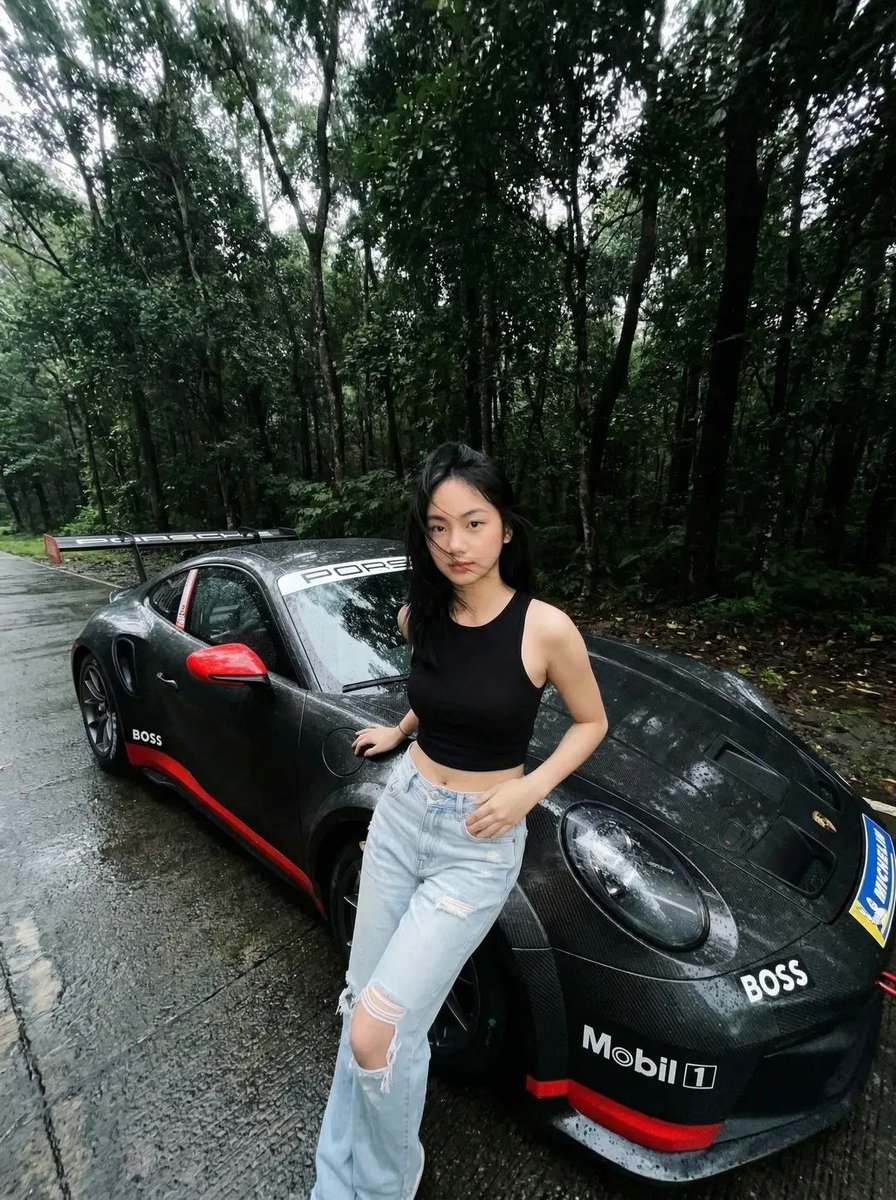 一张电影感、超写实的照片：一位年轻亚洲女性站在雨后湿润的森林道路上，旁边是一辆黑色保时捷跑车。她身穿黑色无袖短款上衣和浅蓝色破洞牛仔裤。她的头…, 图片 1