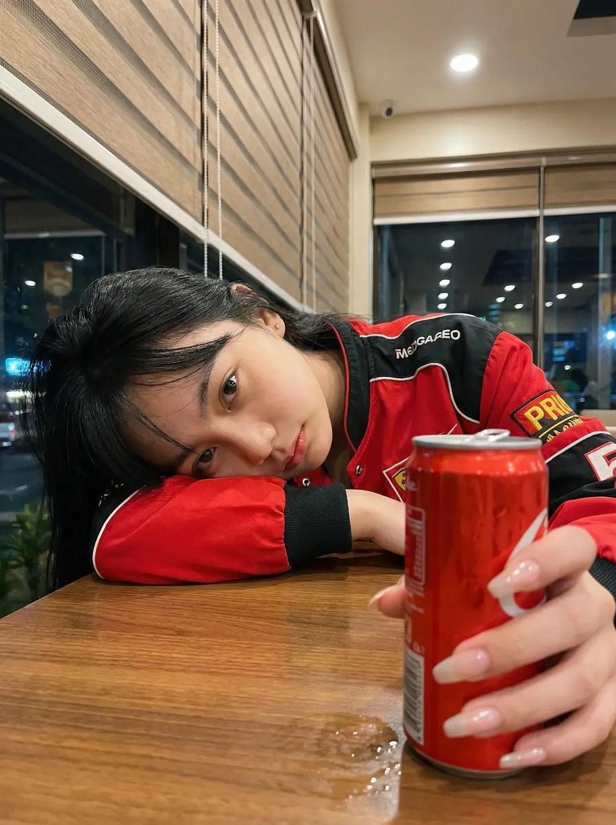 一张超写实的照片：一位年轻女性深夜坐在一间温馨的咖啡馆里，侧着头靠在自己放在木质桌面上的手臂上。她穿着一件红黑配色的赛车风格夹克。表情平静、略…, 图片 1