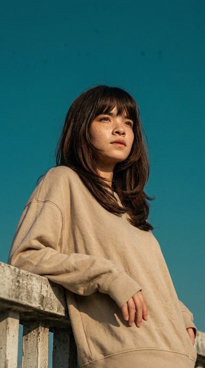 电影感人像照:一位年轻女子倚靠在混凝土栏杆上,身穿宽松米色卫衣,柔和的自然阳光在她脸上投下温暖的色调,背景为清澈的青蓝色天空,淡淡的雀斑,中长…, 图片 1