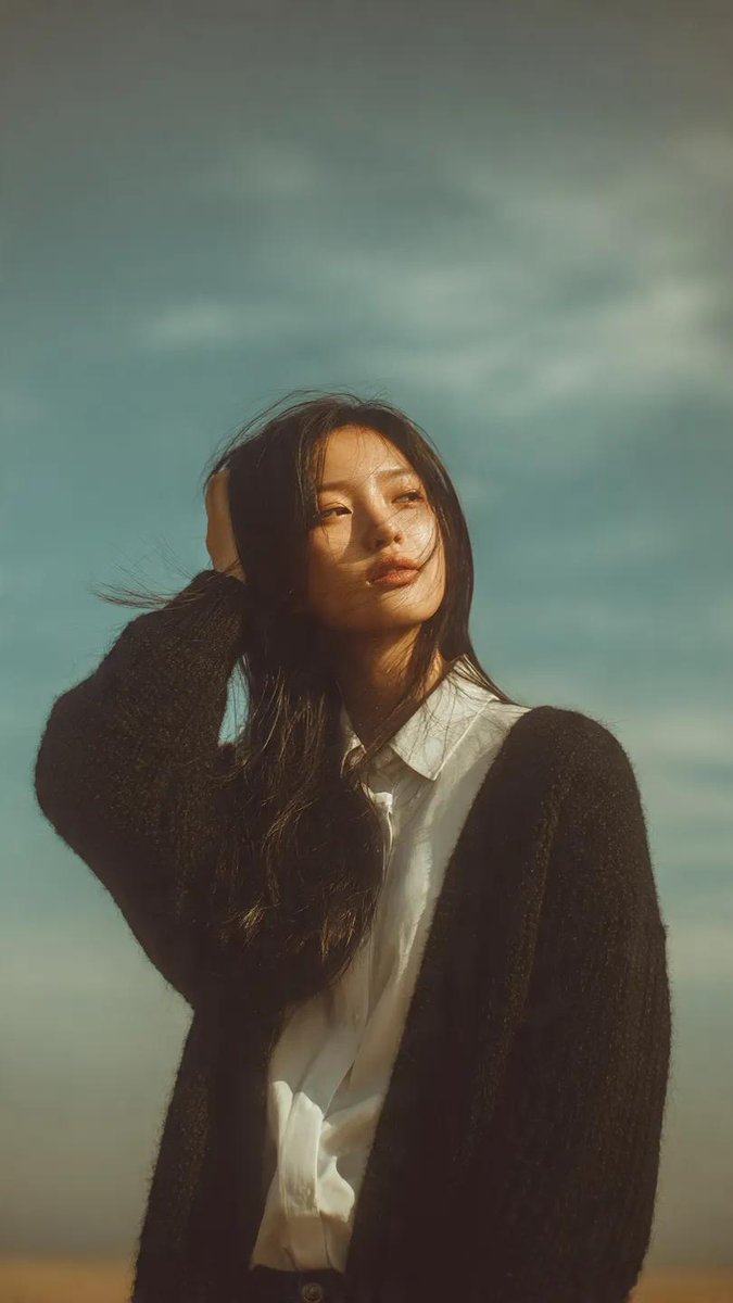 一幅梦幻般的肖像画,一位年轻女子站在户外,头顶是柔和、广袤的蓝天。她那又长又直的黑发在风中轻轻飘扬,脸上带着柔和、怅惘的神情。她身穿一件黑色针…, 图片 3