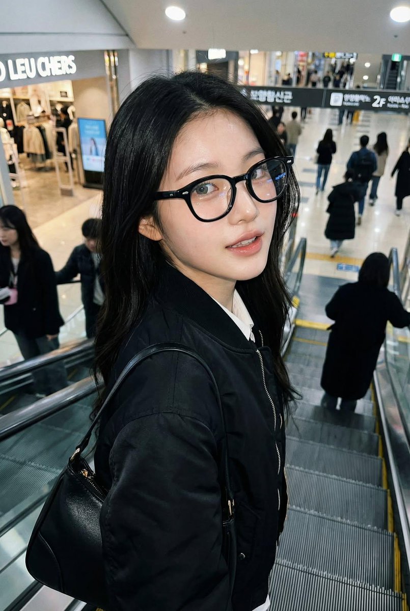 一张像是用 iPhone 拍摄的真实感抓拍照片,画面中是一位女士站在自动扶梯上,面部和身份与参考图完全一致。她戴着黑色玳瑁框眼镜,穿着黑色飞行…, 图片 2