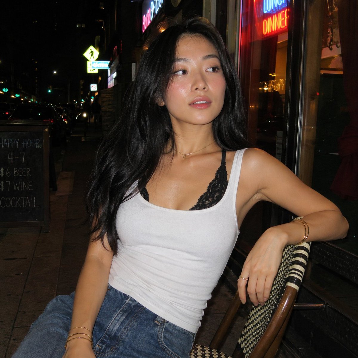 一张夜晚街头的随性人像照,照片中一位年轻亚洲女性随意地坐在一家小型城市餐厅外的藤编咖啡椅上。她有着黑色长发,妆容自然并带有柔和的光泽,身穿白色…, 图片 1