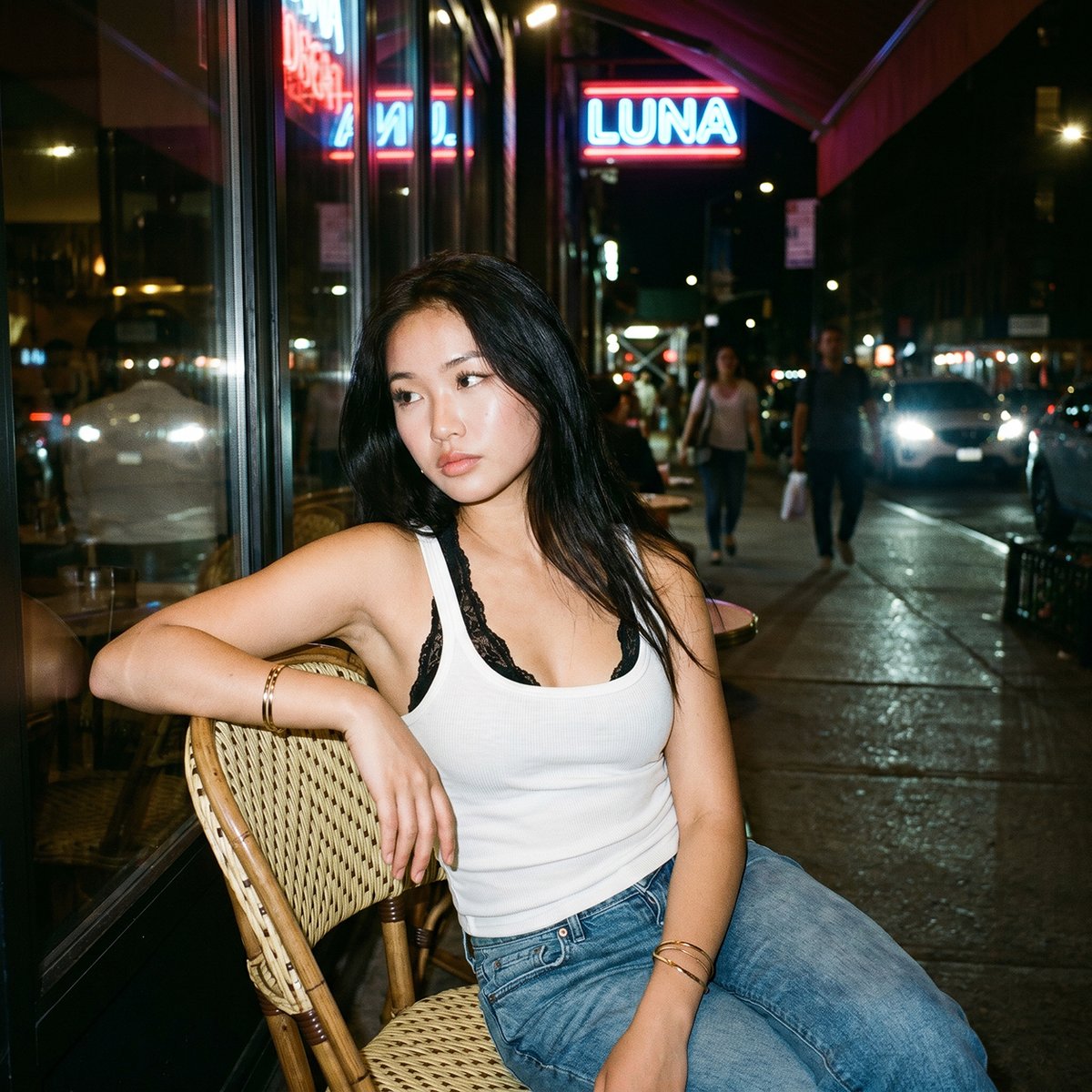 一张夜晚街头的随性人像照,照片中一位年轻亚洲女性随意地坐在一家小型城市餐厅外的藤编咖啡椅上。她有着黑色长发,妆容自然并带有柔和的光泽,身穿白色…, 图片 2