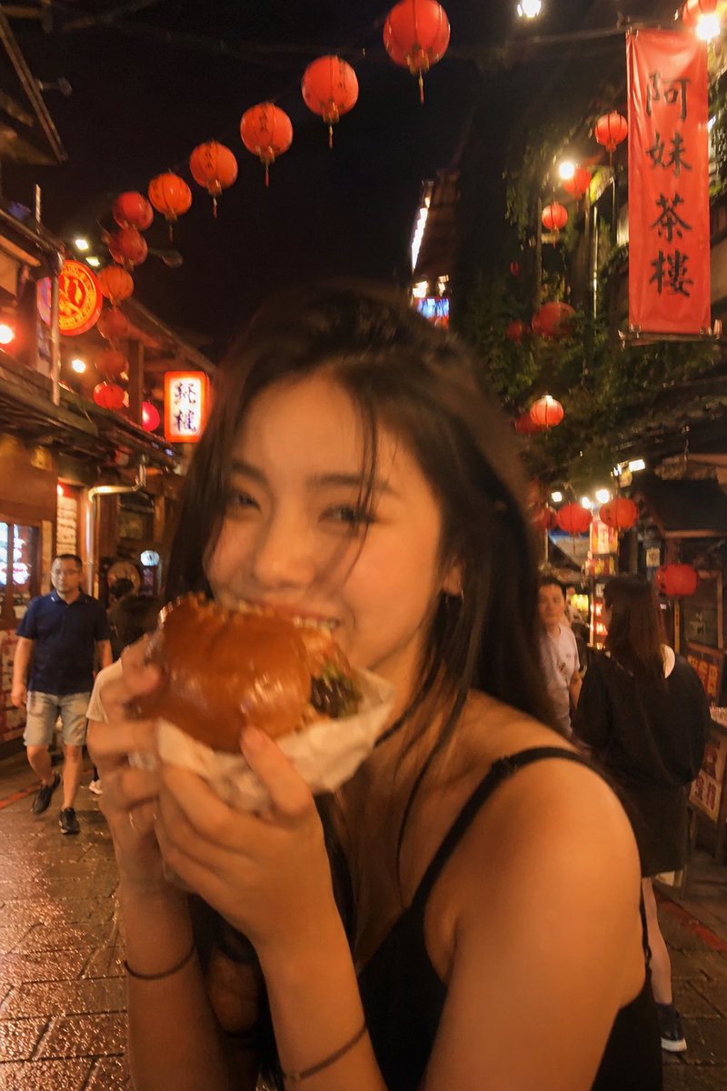失焦的业余快照:一位亚洲超级模特站在夜晚的老台湾街道中央,发光的红灯笼照亮了整个场景。她坐在街边小吃桌前,吃着一个多汁的大汉堡,心情愉悦。暖色…, 图片 1