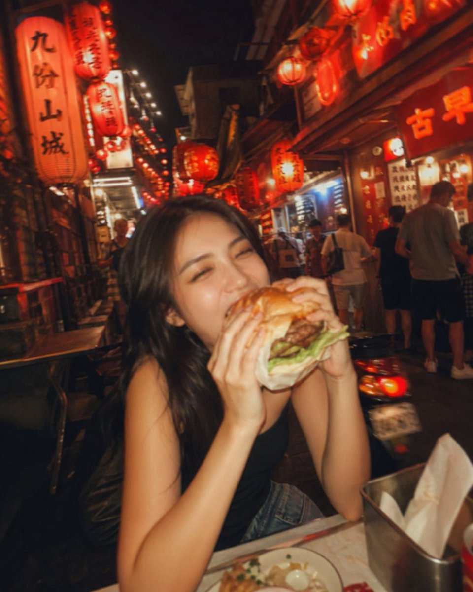 失焦的业余快照:一位亚洲超级模特站在夜晚的老台湾街道中央,发光的红灯笼照亮了整个场景。她坐在街边小吃桌前,吃着一个多汁的大汉堡,心情愉悦。暖色…, 图片 2