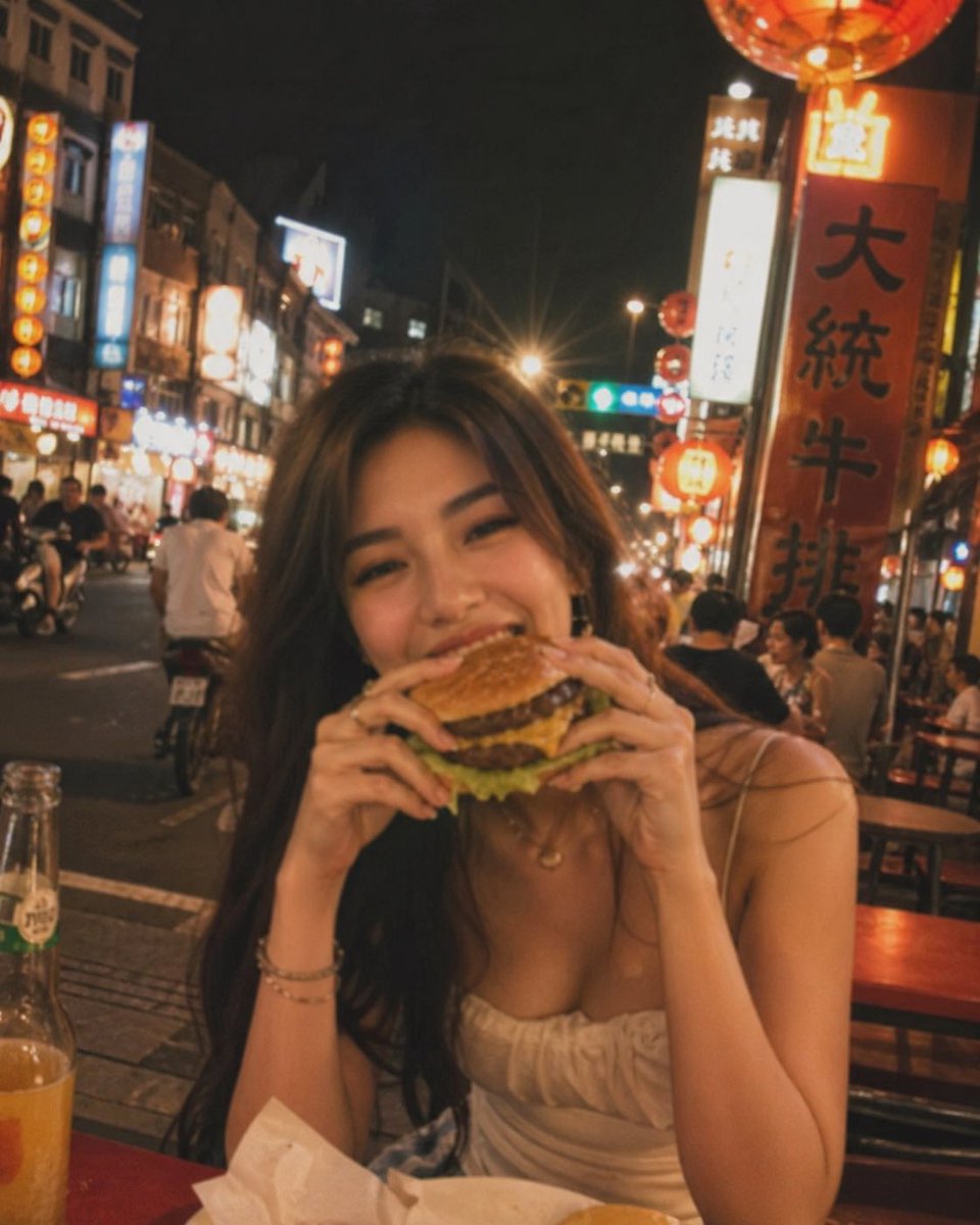 失焦的业余快照:一位亚洲超级模特站在夜晚的老台湾街道中央,发光的红灯笼照亮了整个场景。她坐在街边小吃桌前,吃着一个多汁的大汉堡,心情愉悦。暖色…, 图片 3
