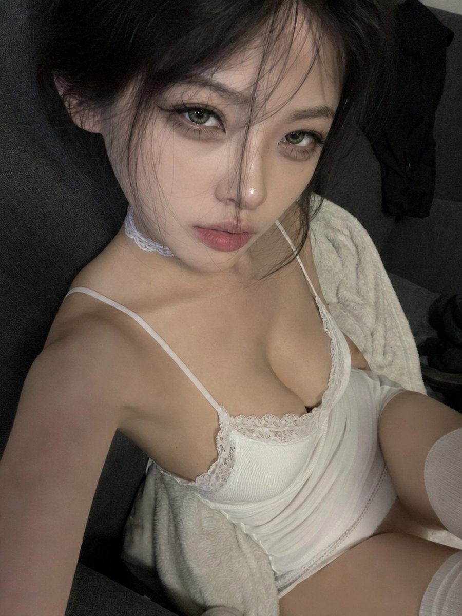 一张近距离高角度的智能手机自拍照，画面中一位年轻女子在夜晚室内斜倚着一张深灰色布艺沙发。手机举在她脸部上方，形成自然的广角畸变，头身比略微放大…, 图片 1