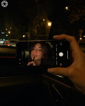 照片拍摄于夜间车内;画面中仅可见女性的一只手和一部iPhone,而年轻女性的面部仅出现在手机屏幕上。相机位于副驾驶一侧,取景框对准挡风玻璃和手…, 图片 1