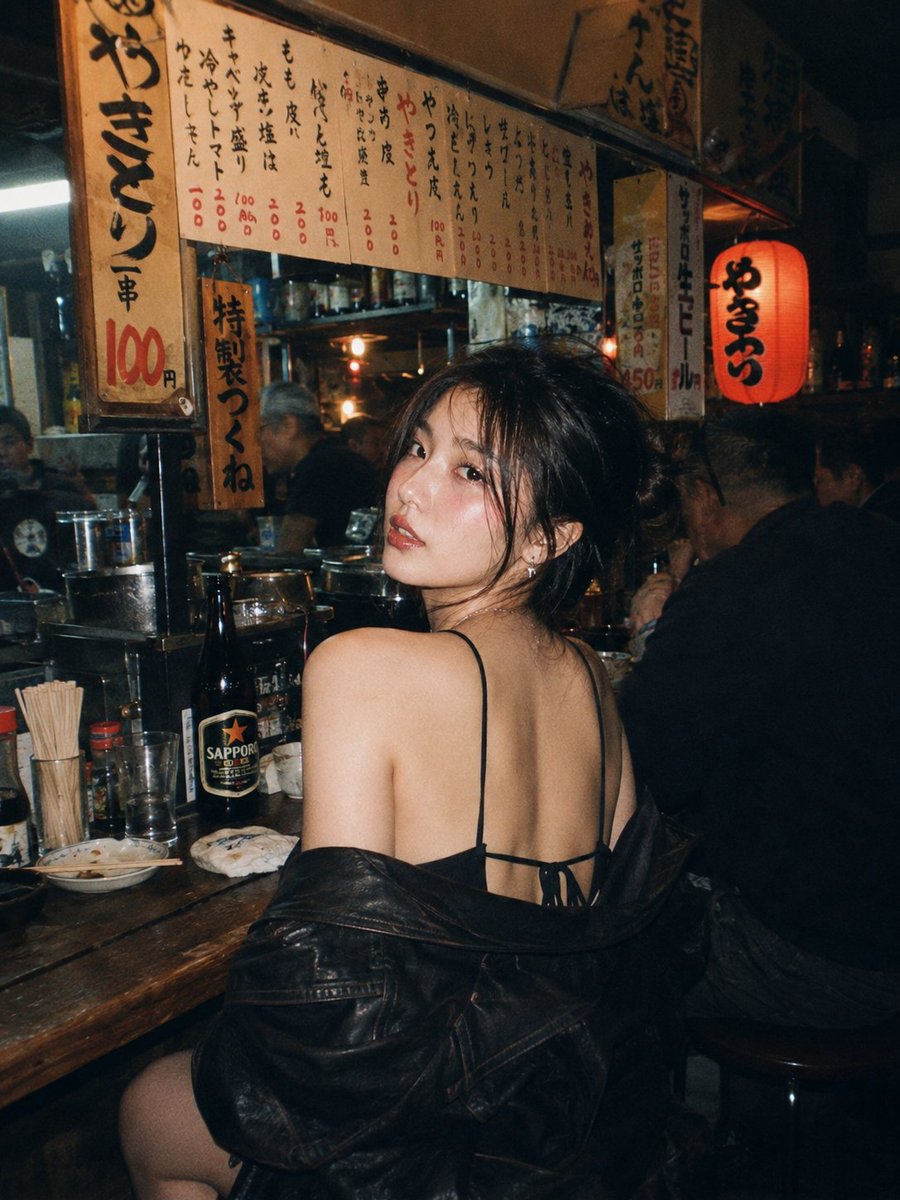 一位美丽的年轻东亚女性，留着深色波浪长卷发，妆容精致，夜晚坐在一家温馨昏暗的日本居酒屋的木质吧台前。她转过头，越过肩膀直视镜头，眼神妩媚诱人。…, 图片 1