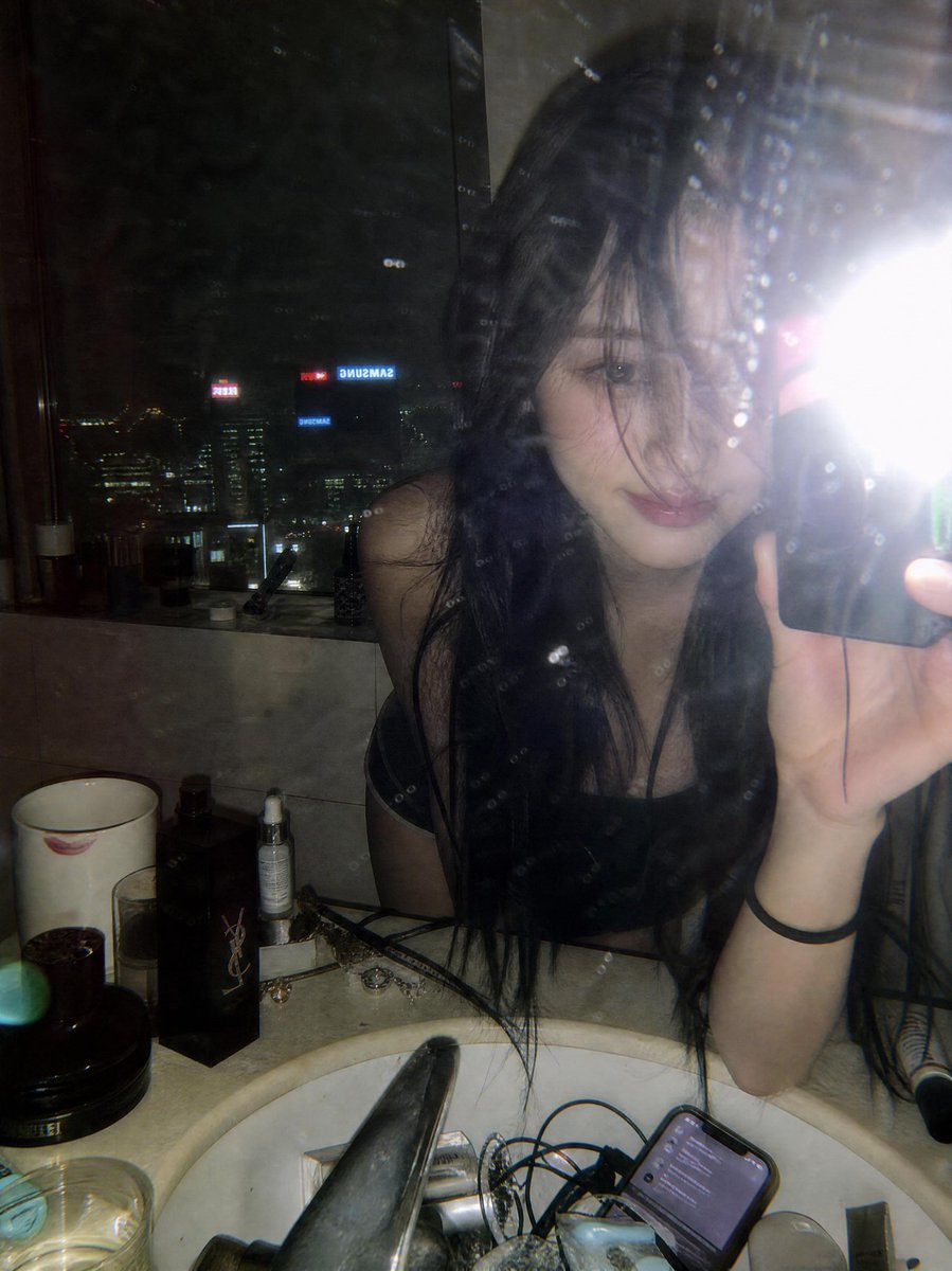 一张在豪华高层公寓浴室中拍摄的深夜情绪化镜子自拍，窗外是灯火辉煌的城市天际线。一位年轻女子留着略凌乱的长黑发，俯身靠在白色大理石洗手台上，零散…, 图片 1