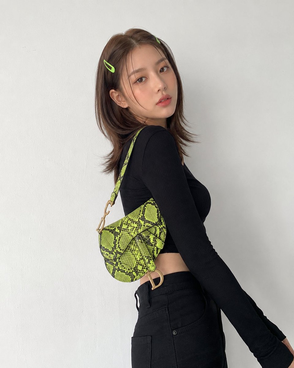 创建一张图片,画面中有一位年轻的韩国女性,留着中长棕色头发,两侧别着青柠绿色的发夹,背靠一面纯白色的墙壁摆姿势,身穿黑色长袖短款上衣、黑色高腰…, 图片 1