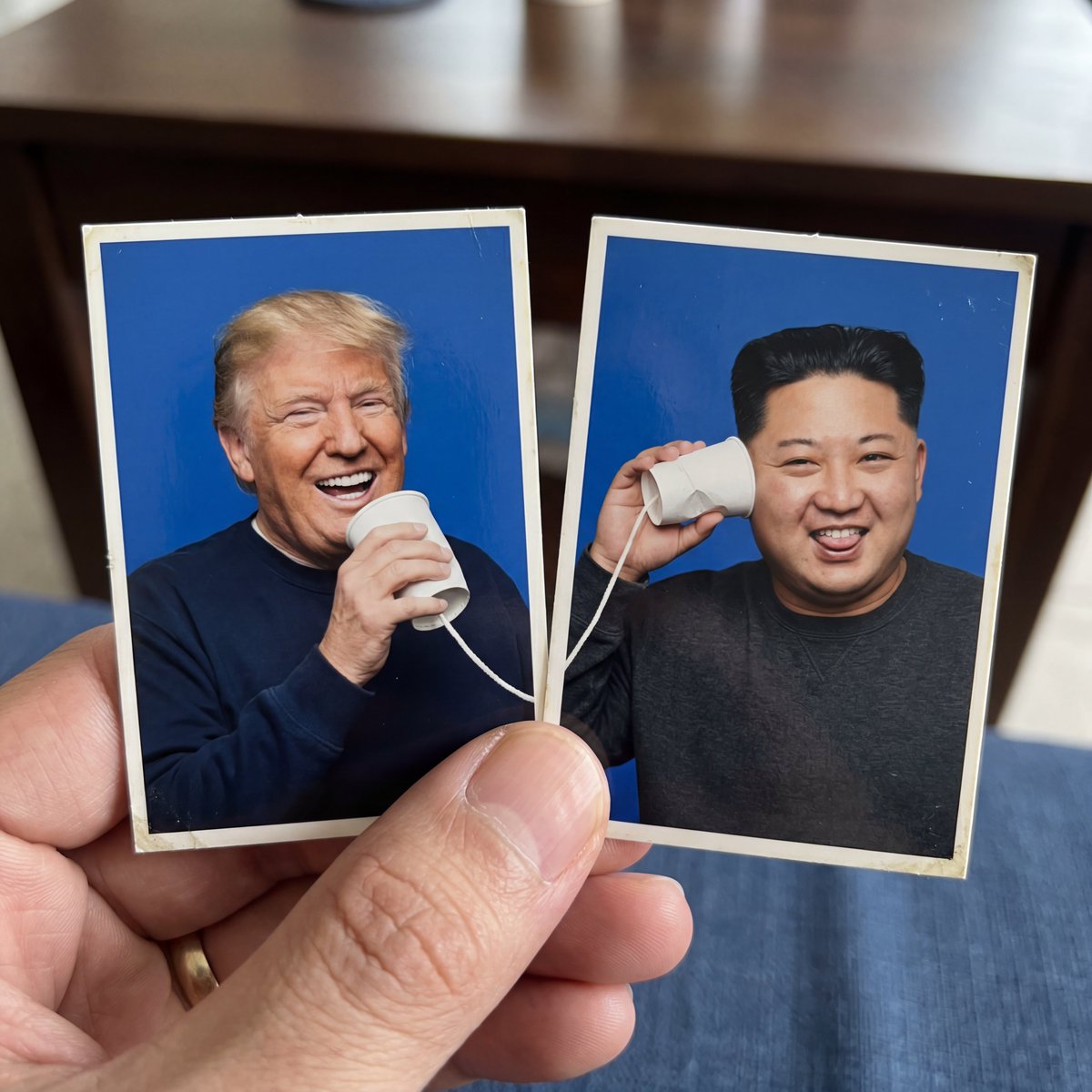 创作一个创意1:1正方形构图,包含两张并排摆放的护照式照片打印件,被一只手的拇指和食指捏住。使用上传的参考图像作为两个拍摄对象,准确保留每个对…, 图片 1