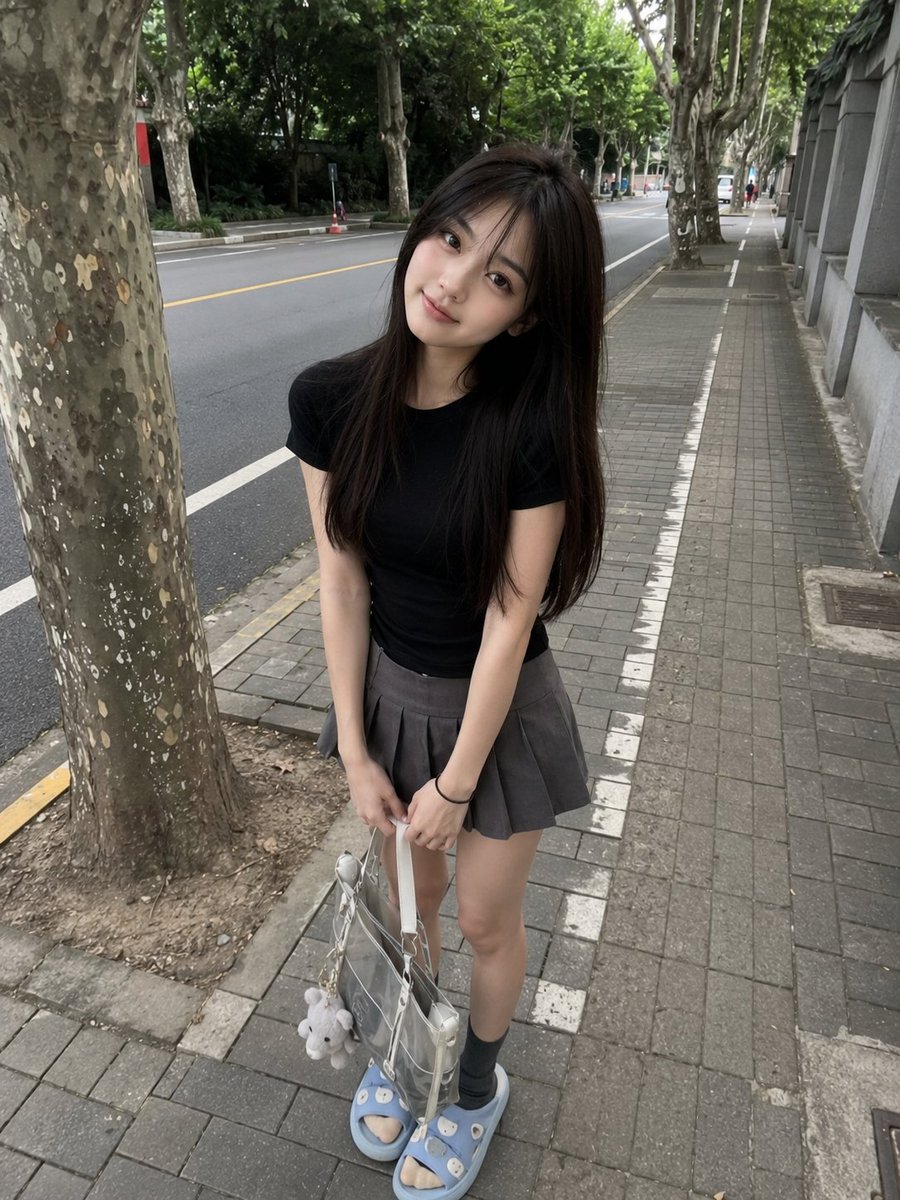 年轻的日本女孩，长直深棕色头发配柔和刘海，身着修身黑色T恤、灰色百褶迷你裙、灰色袜子与浅蓝色拖鞋，随意站在绿树成行的安静道路旁的人行道上。微微…, 图片 2