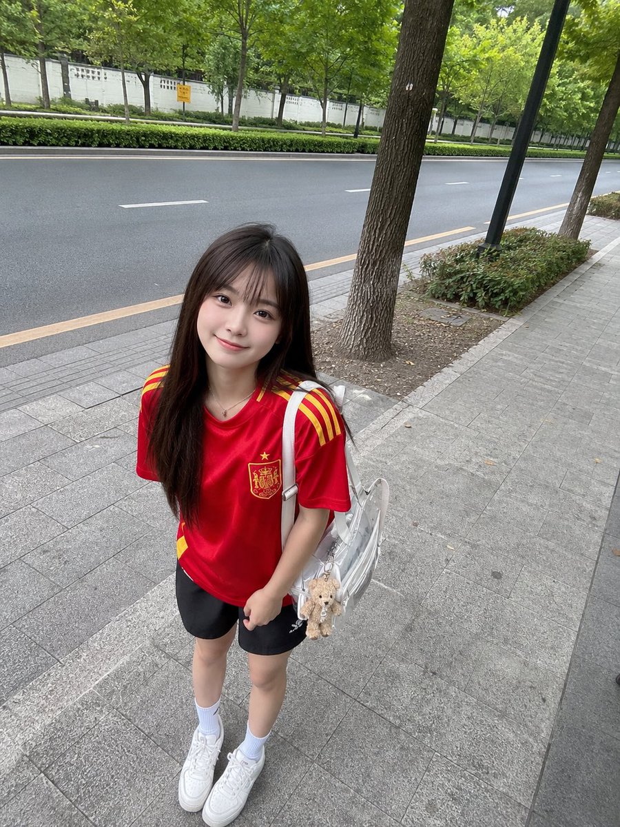 年轻的日本女孩,留着长直的深棕色头发和柔软的刘海,身穿红色西班牙国家足球队球衣、黑色运动短裤、白色中筒袜和白色运动鞋,悠闲地站在绿树成荫的安静…, 图片 2