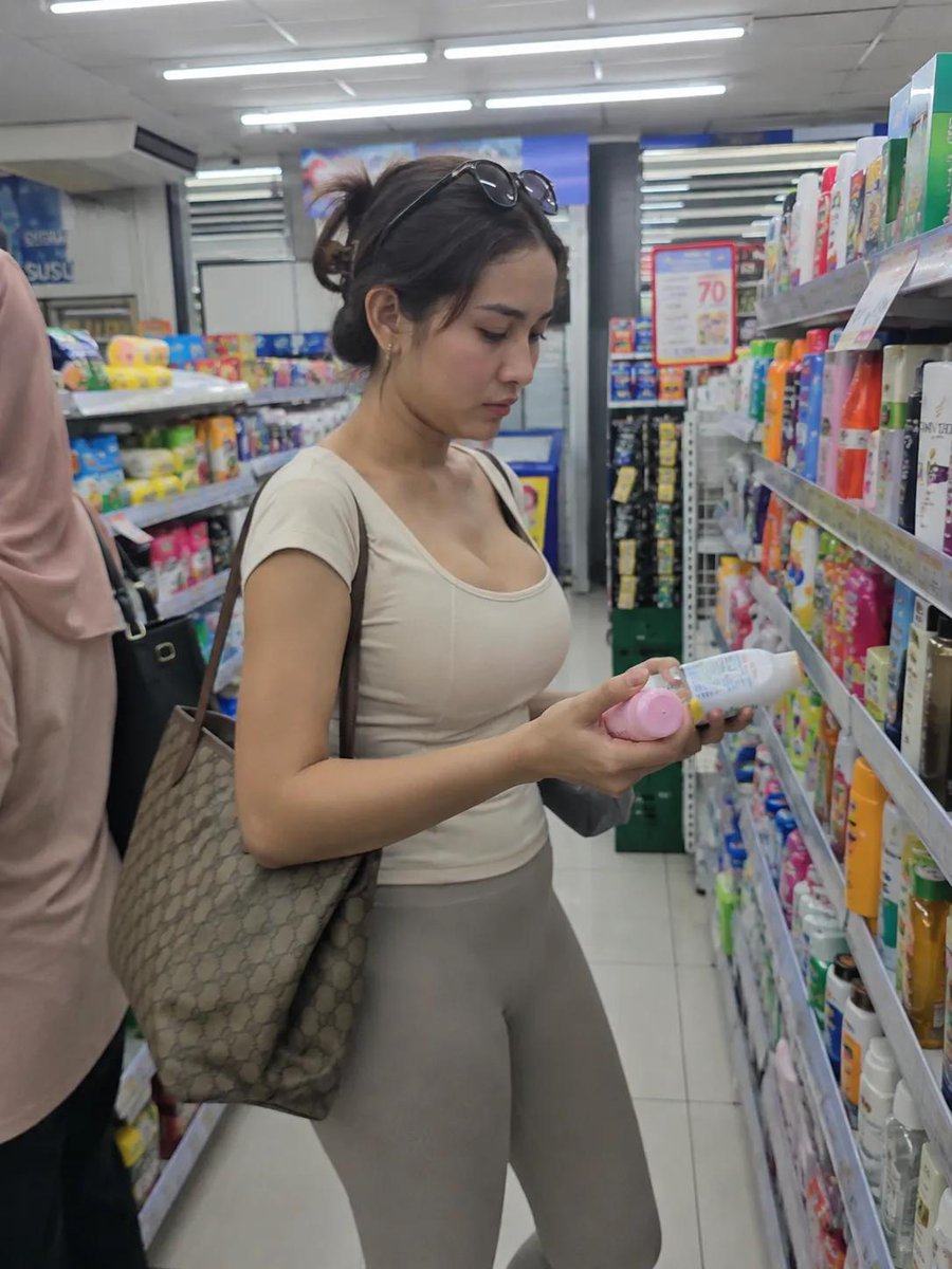 摄影级写实的随性生活照片：一位年轻女性在光线明亮的便利店过道中购物。侧面视角，中全景全身镜头。她站在两排货架之间，货架上摆满了色彩缤纷的个人护…, 图片 1