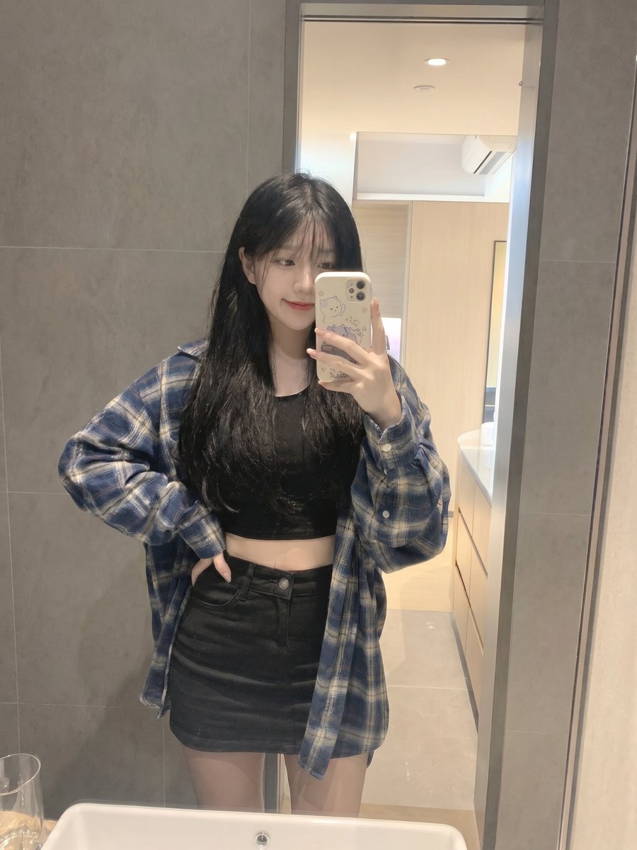 留着长长的黑色直发和柔软稀疏刘海的年轻日本女孩,内穿黑色短款露脐上衣,外穿 oversized 蓝色格纹法兰绒衬衫,搭配黑色迷你短裙。站在浴室…, 图片 1