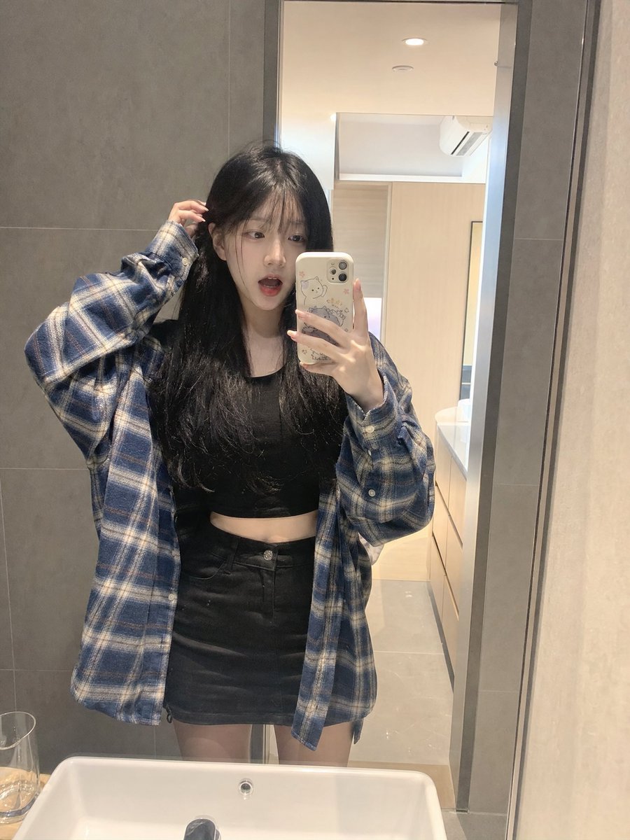 留着长长的黑色直发和柔软稀疏刘海的年轻日本女孩,内穿黑色短款露脐上衣,外穿 oversized 蓝色格纹法兰绒衬衫,搭配黑色迷你短裙。站在浴室…, 图片 2