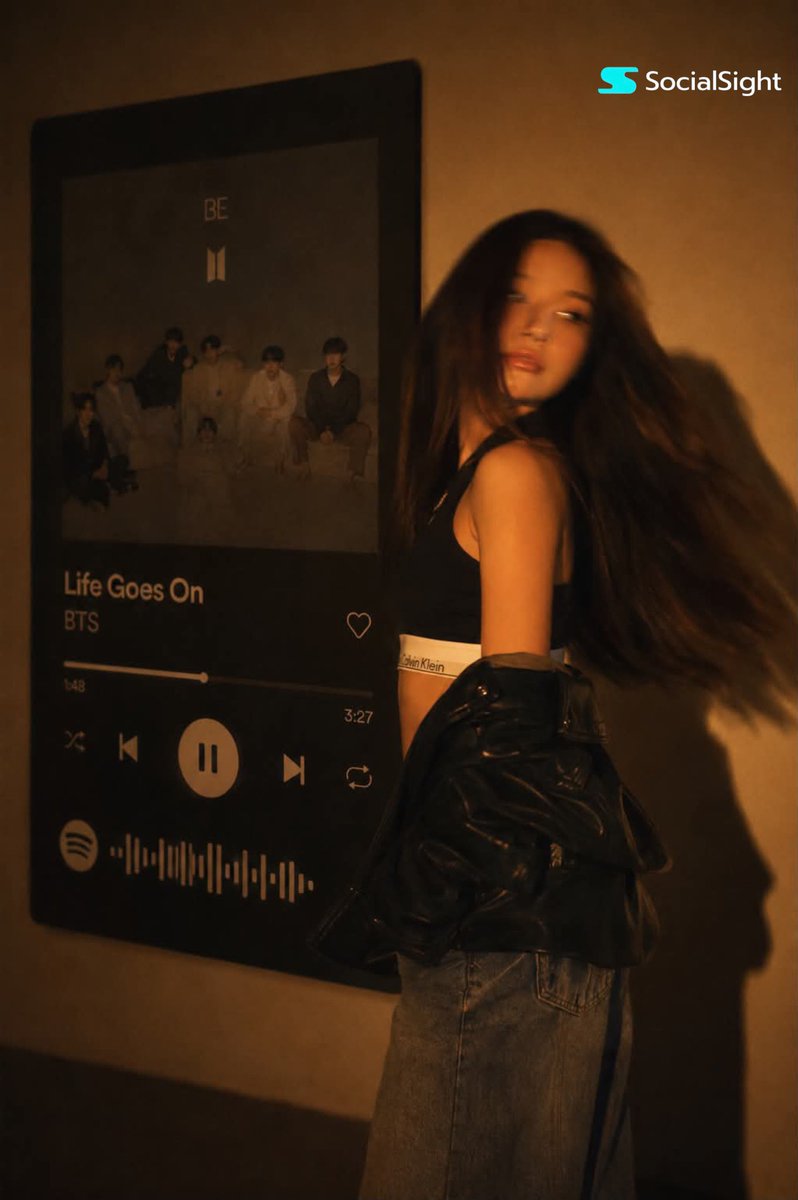 一位发型飘逸的深色长发时尚年轻女子在巨型壁挂Spotify音乐播放器显示屏前摆姿势。温暖的琥珀色电影级灯光布满房间,在墙上投下戏剧性阴影。她身…, 图片 1