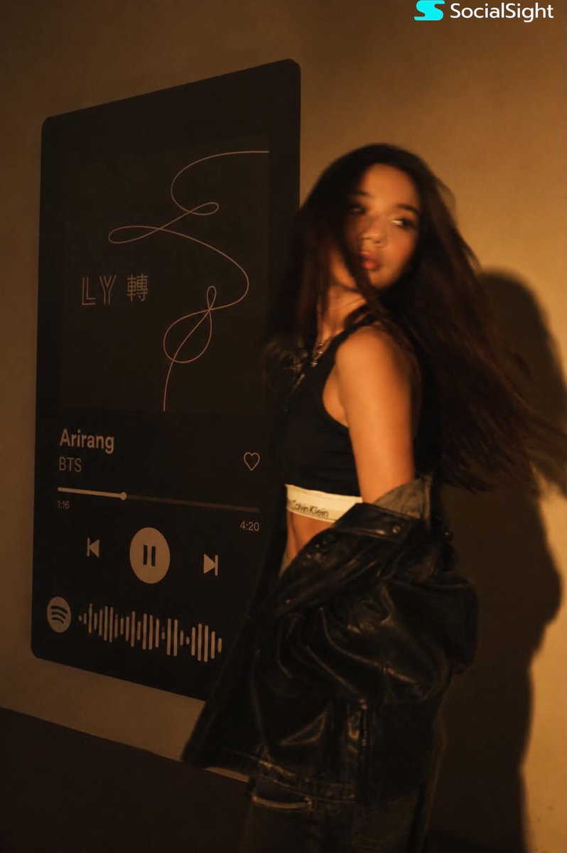 一位发型飘逸的深色长发时尚年轻女子在巨型壁挂Spotify音乐播放器显示屏前摆姿势。温暖的琥珀色电影级灯光布满房间,在墙上投下戏剧性阴影。她身…, 图片 2