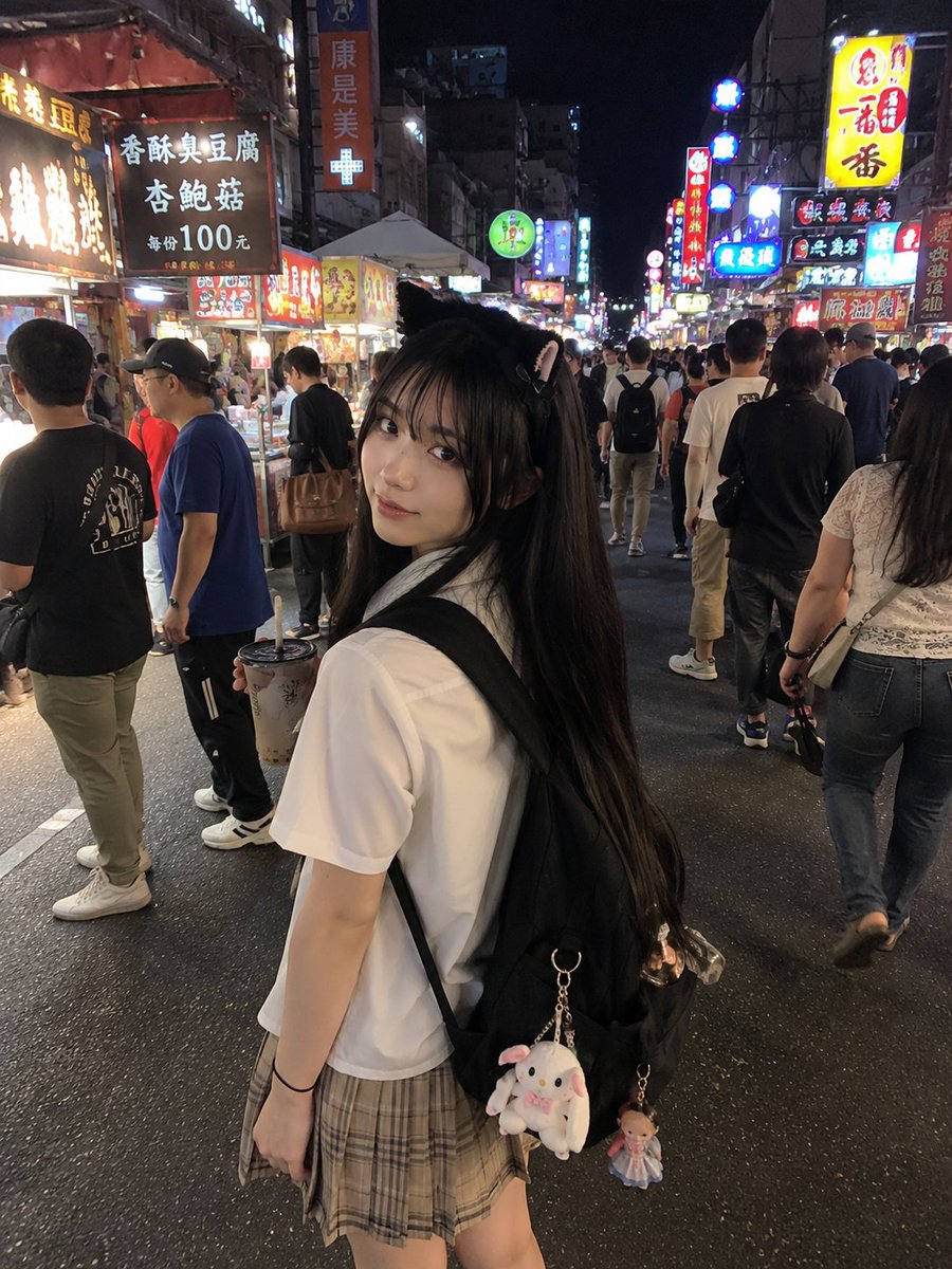 年轻的日本女孩，留着顺直黑色长发和柔软的刘海，戴着可爱的黑色猫耳发箍，身穿白色短袖衬衫、条纹领带和米色格纹百褶裙。她站在一个充满活力的亚洲夜市…, 图片 1