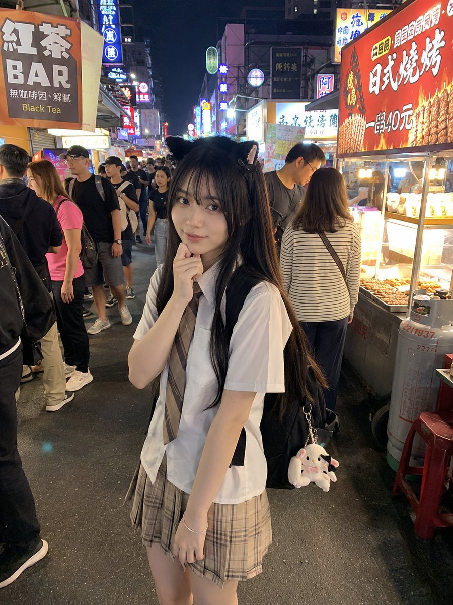 年轻的日本女孩，留着顺直黑色长发和柔软的刘海，戴着可爱的黑色猫耳发箍，身穿白色短袖衬衫、条纹领带和米色格纹百褶裙。她站在一个充满活力的亚洲夜市…, 图片 2