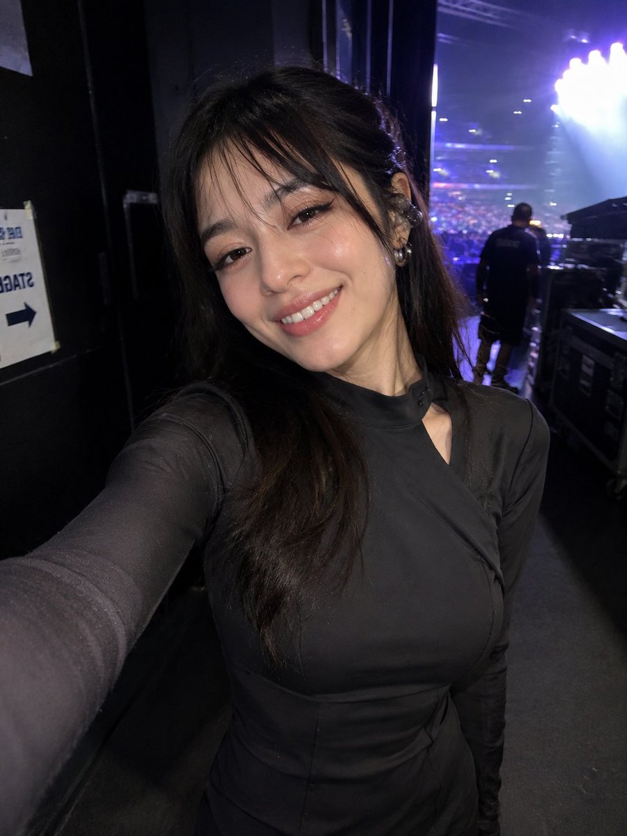 参考图片中同一女性的超写实偶然iPhone前置摄像头自拍,保留完全一致的身份、面部特征、发型和比例。在一场现场演唱会的后台,在她走过走廊时以抓…, 图片 1