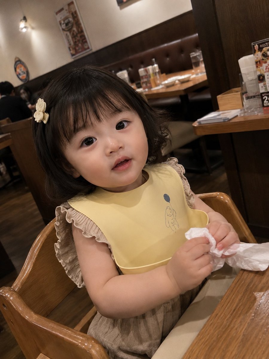 真实感抓拍智能手机照片：一位可爱的女幼儿坐在一家温馨的家庭餐厅的木制高脚椅上。她有着柔软的刘海短发，戴着一个小小的奶白色发夹，眼睛又大又有神，…, 图片 1