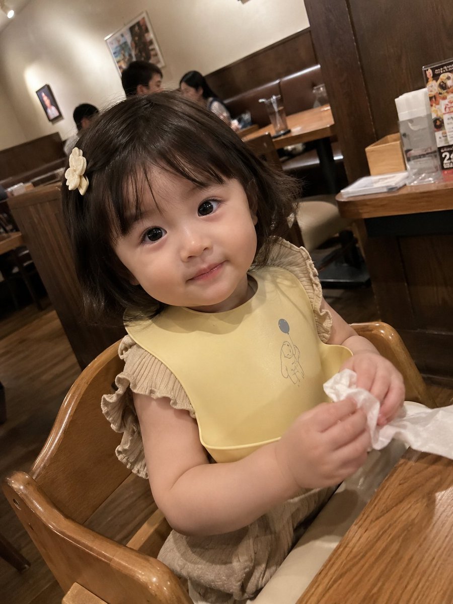 真实感抓拍智能手机照片：一位可爱的女幼儿坐在一家温馨的家庭餐厅的木制高脚椅上。她有着柔软的刘海短发，戴着一个小小的奶白色发夹，眼睛又大又有神，…, 图片 2