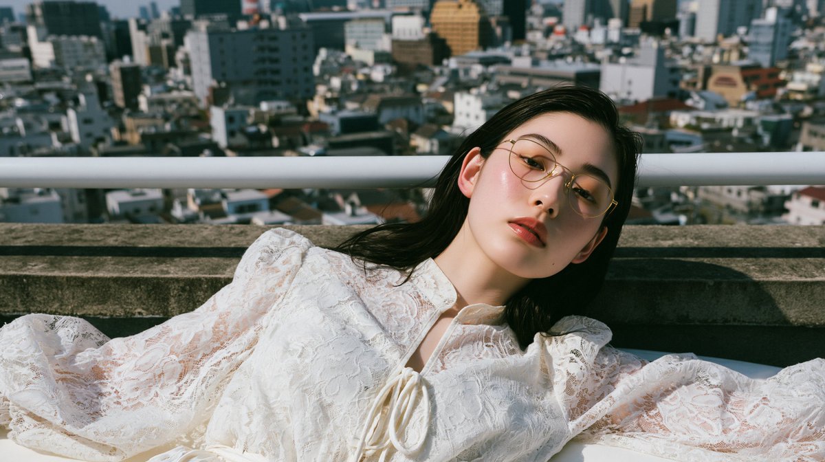 一位惊艳的二十多岁日本或韩国女性，戴着金色金属细框眼镜，身穿白色蕾丝泡泡袖系带领口衬衫，以自然躺卧的姿势斜靠在床沿；头部转向镜头，位于高楼屋顶…, 图片 1