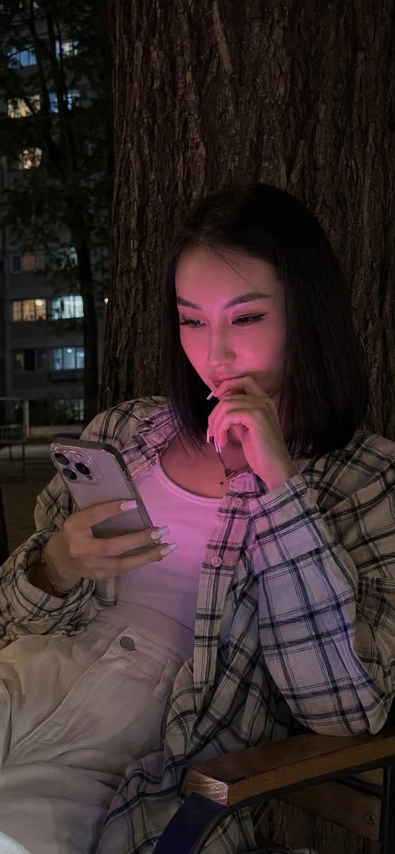 夜晚使用 iPhone 15 Pro 拍摄的写实风格生活照。画面中央是一位年轻女性，留着及腰的黑色长直发。她正饶有兴趣地注视着智能手机，嘴角轻…, 图片 1
