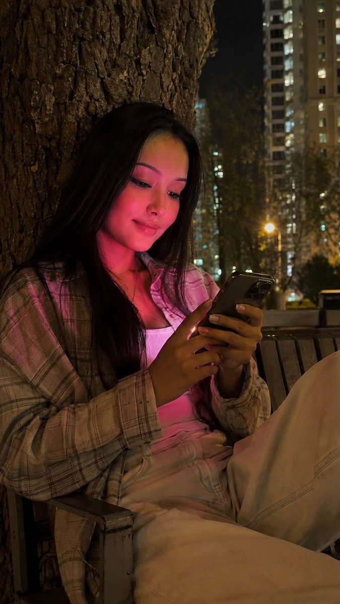 夜晚使用 iPhone 15 Pro 拍摄的写实风格生活照。画面中央是一位年轻女性，留着及腰的黑色长直发。她正饶有兴趣地注视着智能手机，嘴角轻…, 图片 2