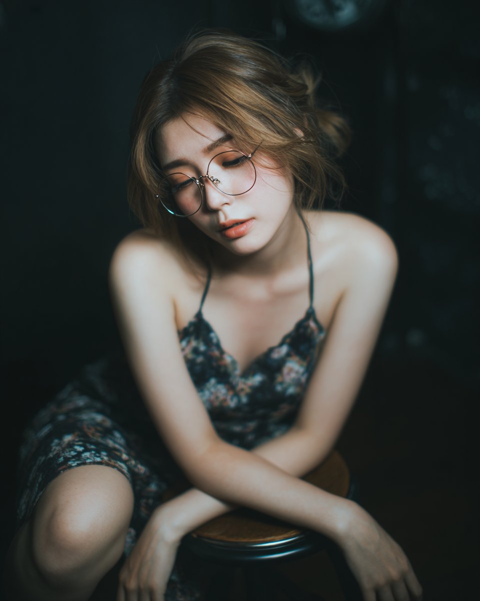 一位二十多岁的迷人日本或韩国女性，戴着金色金属细框眼镜，身穿长裙，以坐姿安排坐在吧台凳上，一膝屈起、另一条腿自然放松；头部低垂朝双手方向，身处…, 图片 1