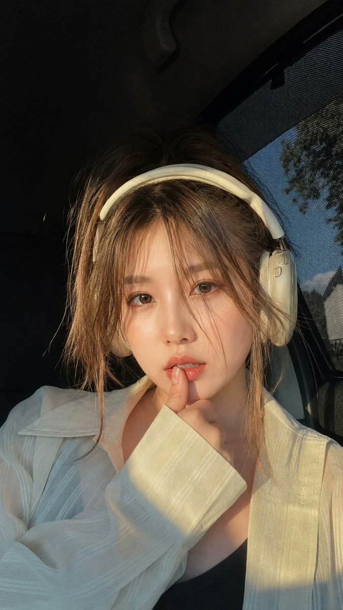 黄金时段坐在豪华轿车内的美丽年轻女子的超写实特写肖像,佩戴优雅的米白色头戴式耳机。柔和的自然阳光从侧面车窗洒入,在她的脸庞和宽松的白色衬衫上投…, 图片 1