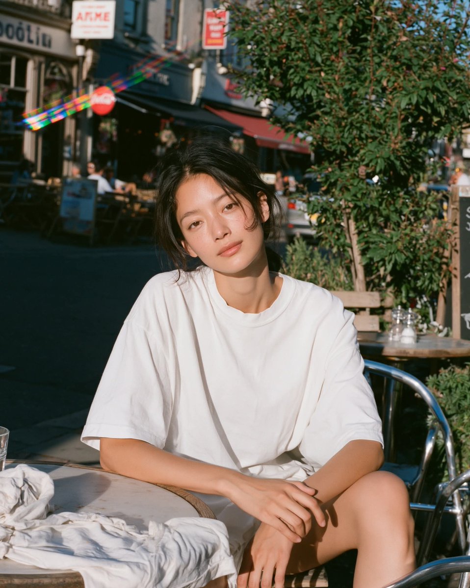 温暖阳光下的胶片人像,年轻女性放松地坐在小咖啡馆露台桌旁沐浴在明亮的金色午后阳光下,目光平静柔和、双眼睁开,身穿超大号白色T恤,身后是清晰可辨…, 图片 1