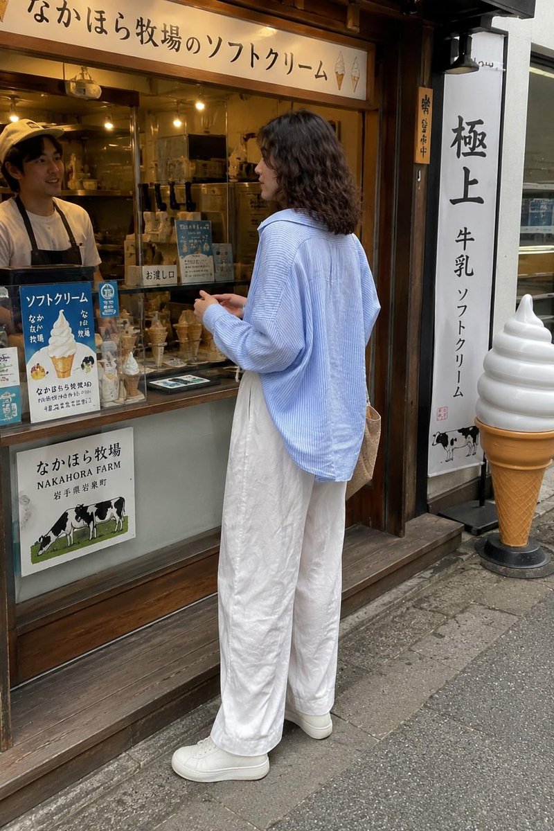 一张抓拍的旅行照片:一位年轻女子站在一家传统日式软冰淇淋店外,回眸对镜头微笑。她留着齐肩深色波浪卷发,身穿宽松的浅蓝色条纹纽扣衬衫、宽松的白色…, 图片 2