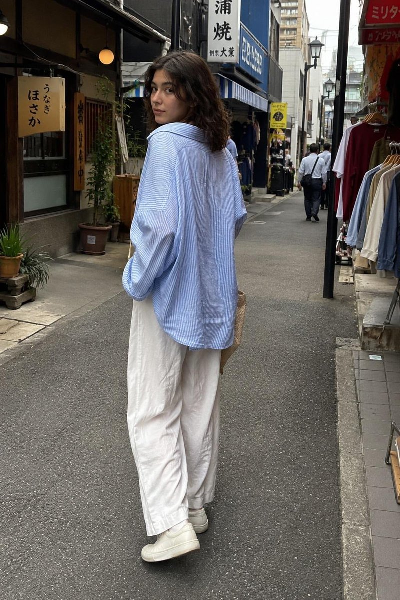 一张抓拍的旅行照片:一位年轻女子站在一家传统日式软冰淇淋店外,回眸对镜头微笑。她留着齐肩深色波浪卷发,身穿宽松的浅蓝色条纹纽扣衬衫、宽松的白色…, 图片 3