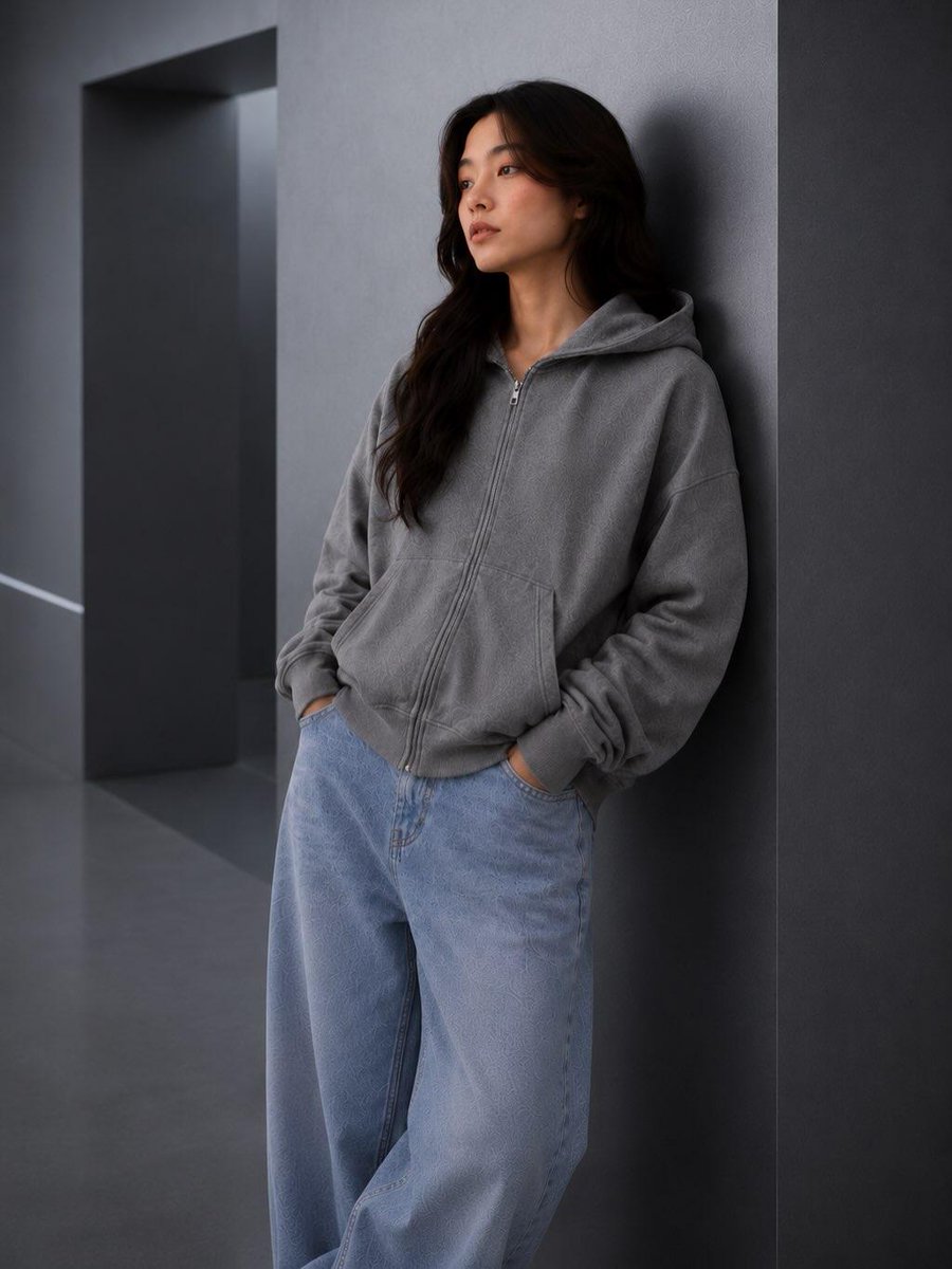 极简时尚编辑人像:一位年轻女性随意倚靠在一面光滑的炭灰色建筑墙面上,身穿水洗灰色宽松拉链连帽衫、浅色水洗宽松牛仔裤和白色运动鞋,双手插兜,姿态…, 图片 1