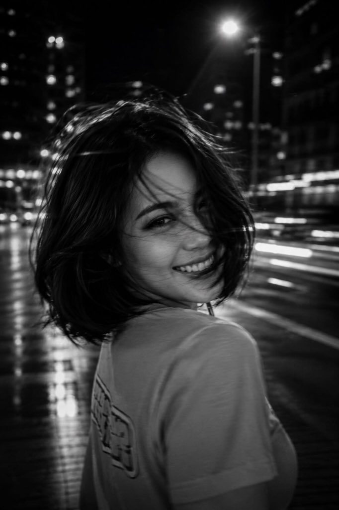 A black-and-white cinematic photograph of a young woman at night in the city, s…