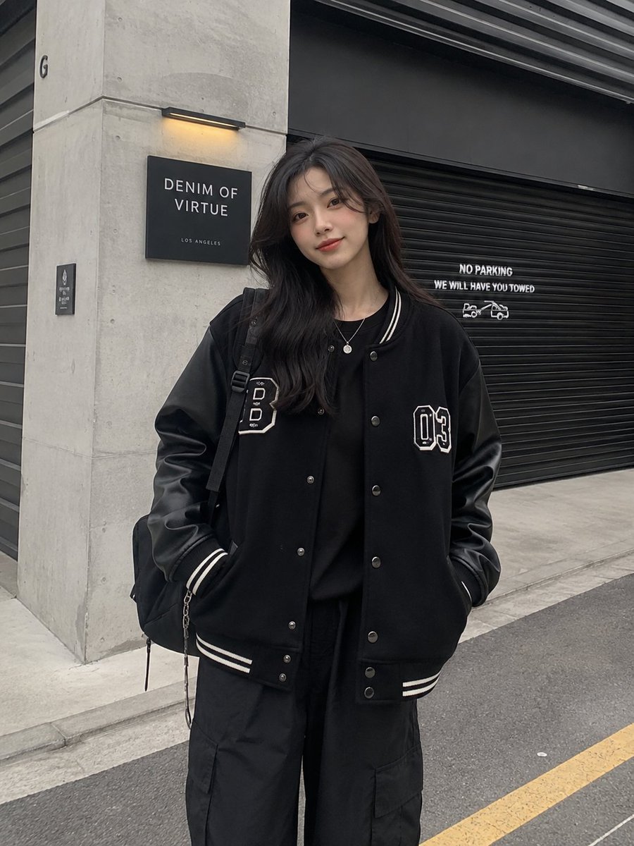一张潮流街头服饰时尚人像照:一位时尚年轻女性自信地站在一座现代工业建筑前,建筑拥有混凝土墙面和一扇黑色金属卷帘门。她身穿一件超大版型的黑色棒球…, 图片 1