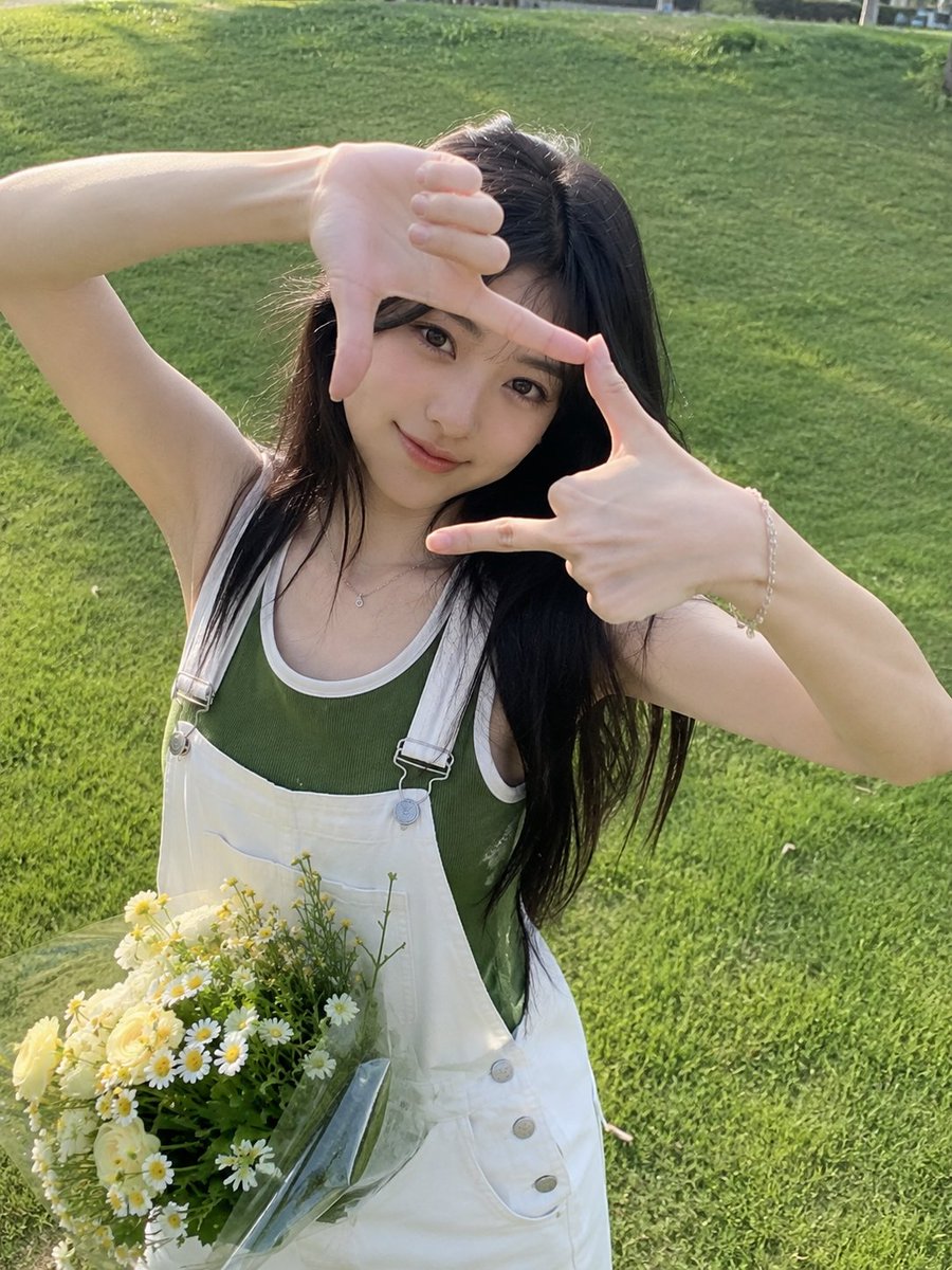 一张迷人的户外人像照,一位年轻女子站在郁郁葱葱的绿色草坪上,直视镜头,双手俏皮地在脸部周围比划出一个长方形相框。她有着一头长而深色、柔和波浪卷…, 图片 1