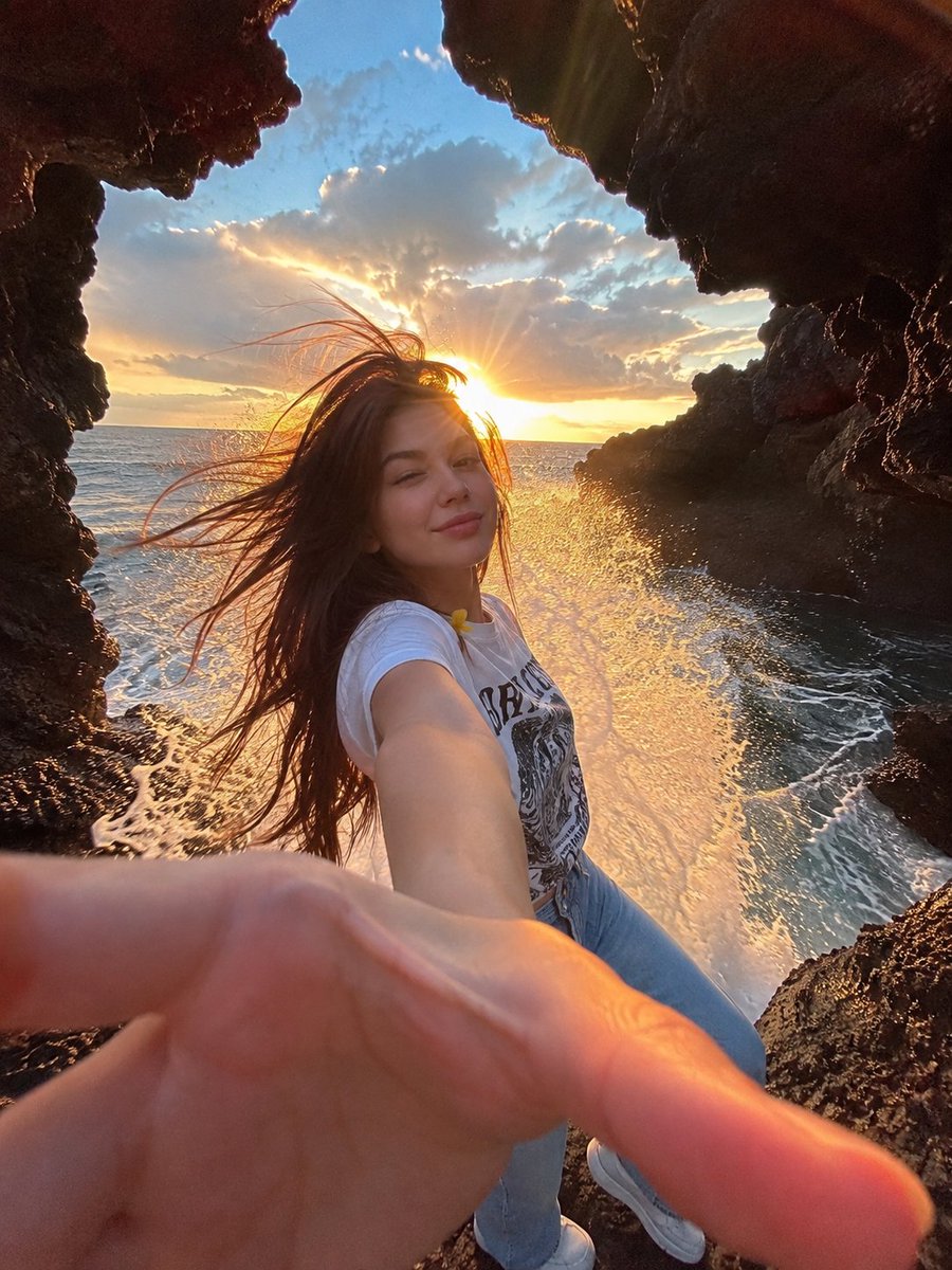一幅电影感十足的海边人像照:一位年轻女子站在天然海洞内的黑色火山岩上,洞口粗犷的岩石形成天然画框。她一头深色长发在海风中戏剧性地飘扬,对着镜头…, 图片 2