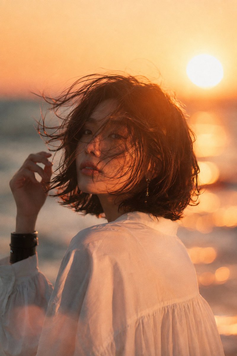 海滩日落中的美丽年轻亚洲女性,中景电影肖像。深色短发波波头在海风中飘扬,头部微向左倾,手正在整理头发。甜美浪漫的瞬间。精致优雅的耳饰,简约宽松…, 图片 2