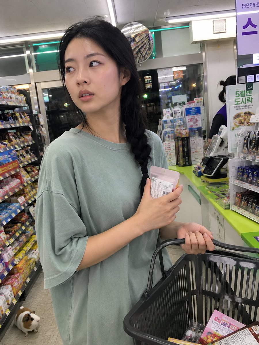 用iPhone前置摄像头意外抓拍的快照，拍摄于一家小型社区便利店内部。一位年轻的韩国女性站在收银台前排队，一边随意地用手抹去额头上的一丝汗珠，…, 图片 2