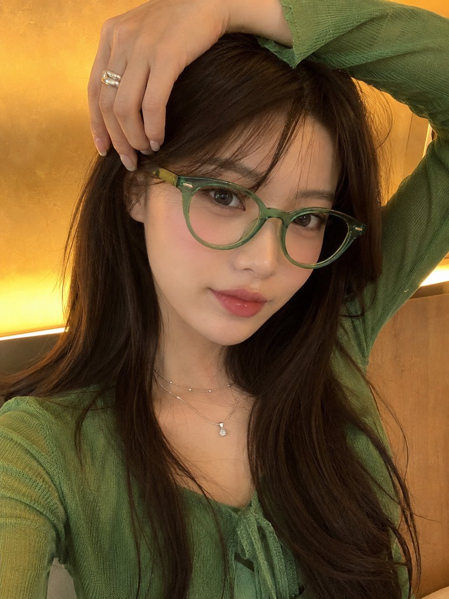 一张高品质的竖版人像照，主体是一位年轻女性，留着长直深棕色头发，戴着时尚的半透明翡翠绿猫眼眼镜，身穿鲜艳的绿色服装。她在室内摆姿势，一只手自然…, 图片 1
