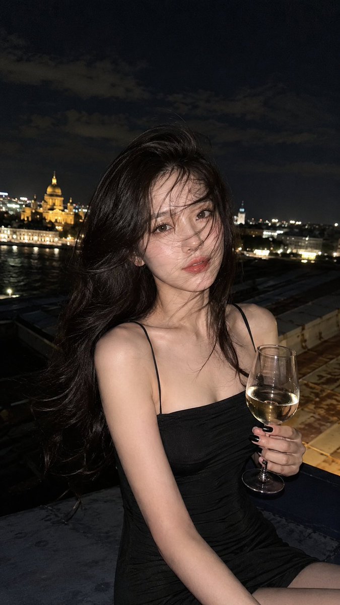 使用参考图像。圣彼得堡屋顶上的半身夜景肖像,一位女性手持一杯白葡萄酒。她身穿修身的黑色吊带连衣裙,露出肩膀,指甲剪短涂成黑色。蓬松有光泽的长发…, 图片 2